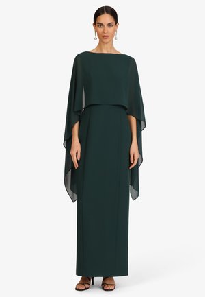 Frau trägt ein langes dunkelgrünes Kleid mit transparenten weiten Ärmeln und schwarzen Riemchen-High Heels, steht vor einem schlichten weißen Hintergrund.