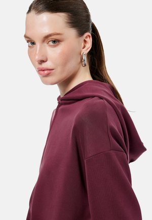 Vrouw met naar achteren gebonden haar, die grote zilveren oorringen en een donkerrode hoodie draagt, licht naar de camera kijkend tegen een effen achtergrond.