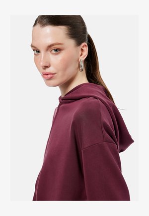 Kobieta z włosami związanymi do tyłu, nosząca duże srebrne kolczyki koła oraz bordową bluzę z kapturem, delikatnie patrząca w stronę aparatu na jednolitym tle.