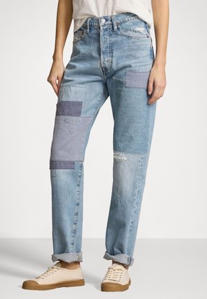 Jean en denim patchwork bleu clair avec revers roulés, porté avec des baskets beige et une chemise blanche rentrée.