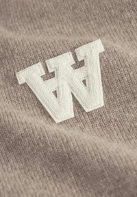 Tan strikket stof med et stort, hvidt broderet bogstav "W" med en tekstureret overflade. Blød tekstur med tydelige syning detaljer.