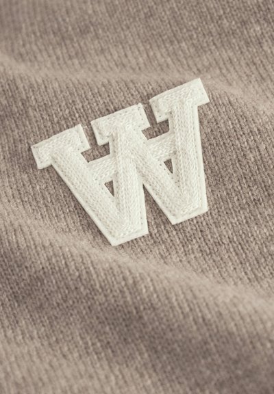 Tan knit fabric featuring a large, white embroidered letter "W" with a textured finish. Soft texture with distinct stitching details.