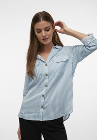 Camicia button-up a righe blu chiaro con colletto, due tasche frontali e bottoni marroni. Il tessuto appare liscio e ha una vestibilità comoda.