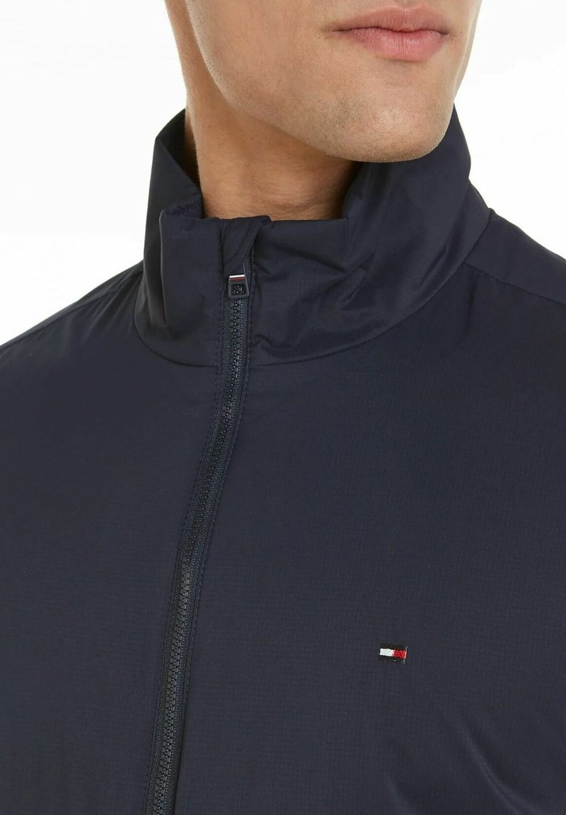 Giacca blu navy con cerniera e collo alto, caratterizzata da un tessuto strutturato e un piccolo logo sul petto a sinistra. Design elegante e moderno.