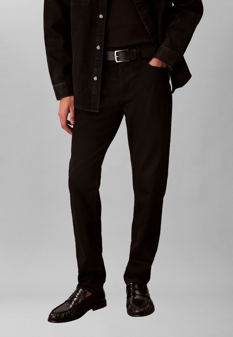 Άνδρας που φοράει μαύρο τζιν, μαύρη ζώνη, μαύρο πουκάμισο και  μαύρα δερμάτινα μοκασίνια, με το ένα χέρι στην τσέπη, στέκεται μπροστά σε απλό γκρι φόντο.