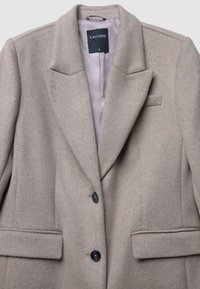 Cappotto beige in misto lana con colletto a risvolto, due tasche frontali, tre bottoni neri e una fodera interna liscia. Etichetta del marchio visibile all'interno.
