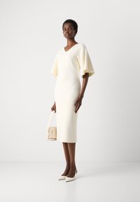 Cream-colored fitted dress with short, flared sleeves. Smooth texture, V-neck design. Paired with a light handbag and pointed heels.