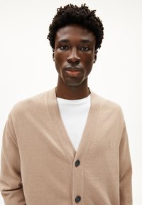 Beige cardigan met een V-hals ontwerp, zachte textuur, drie zwarte knopen en subtiele stikdetails, gedragen boven een wit overhemd.