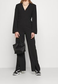 Mono negro con mangas largas, escote en V, bolsillos laterales y dobladillos abiertos. Combinado con zapatillas negras y un pequeño bolso negro.