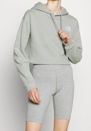 Persoon die een lichtgrijze cropped hoodie en bijpassende grijze high-waisted sportfietsbroek draagt, staand tegen een witte achtergrond.