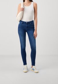 Jeans de denim de corte ajustado y de color azul oscuro con cintura alta, diseño de cinco bolsillos y un acabado suave, combinados con una camiseta sin mangas blanca y zapatillas deportivas.