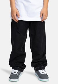 Pantalones de mezclilla negra con costuras blancas, combinados con zapatillas grises que tienen cordones blancos y acentos en teal, llevados con una camiseta blanca.