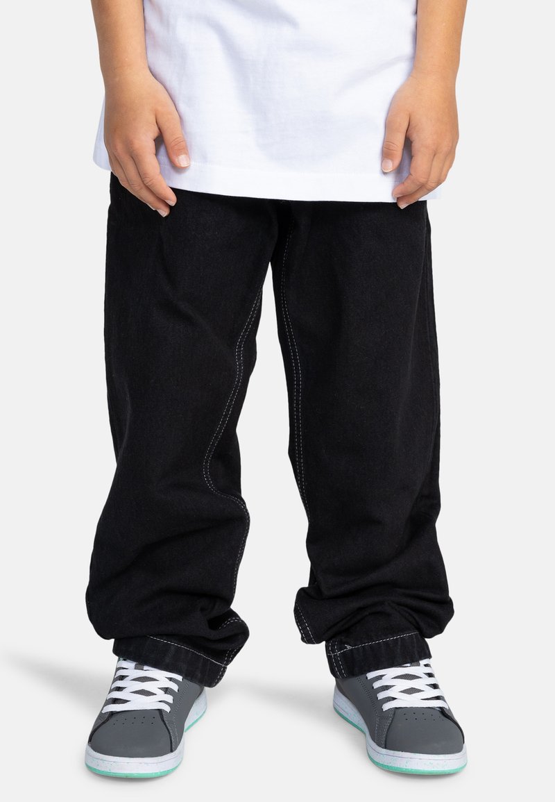 Pantalones de mezclilla negra con costuras blancas, combinados con zapatillas grises que tienen cordones blancos y acentos en teal, llevados con una camiseta blanca.