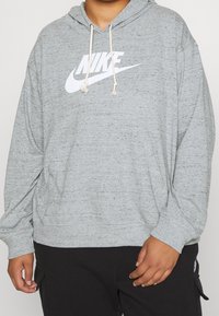Person som bär en grå Nike-hoodie med vitt logotyp och dragsko, ihop med svarta byxor med sidofickor.