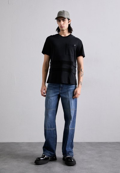 Fekete, rövid ujjú ing zsebbel és texturált vízszintes csíkokkal, közép-kék foltvarrott farmernadrággal és vastag fekete cipővel párosítva.