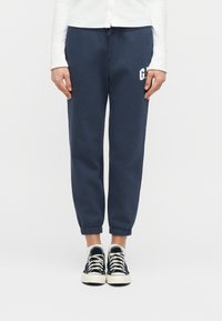 Άτομο που φοράει σκούρο μπλε παντελόνι φόρμας με ένα μικρό λευκό λογότυπο, μαύρα και λευκά αθλητικά παπούτσια και λευκές κάλτσες, στέκεται μπροστά σε ένα απλό λευκό φόντο.