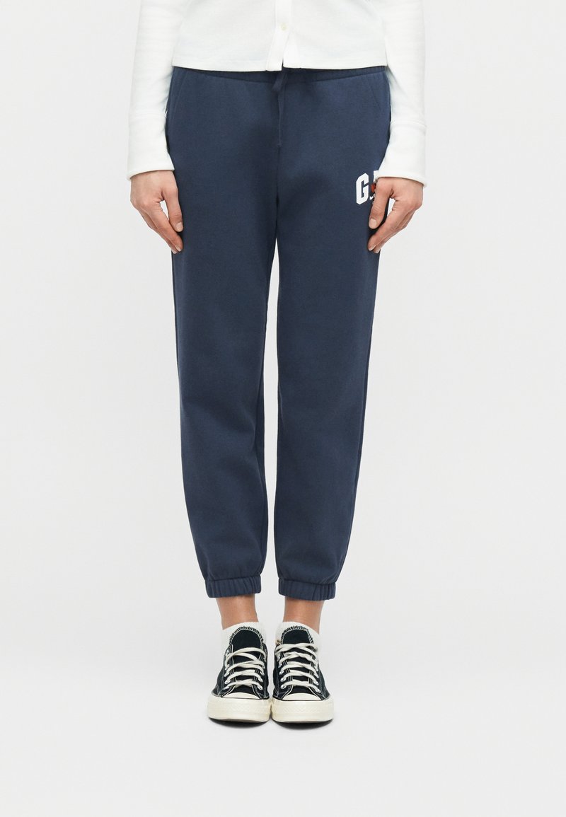 Άτομο που φοράει σκούρο μπλε παντελόνι φόρμας με ένα μικρό λευκό λογότυπο, μαύρα και λευκά αθλητικά παπούτσια και λευκές κάλτσες, στέκεται μπροστά σε ένα απλό λευκό φόντο.