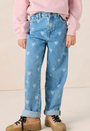 Child wearing light blue denim jeans with heart patterns, rolled cuffs, beige platform sneakers, and a pink long-sleeve top.