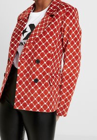 Röd kordfläskjacka med diamantmönster i vitt, dubbelknäppt med svarta knappar och sidofickor, kombinerad med en vit grafisk t-shirt.