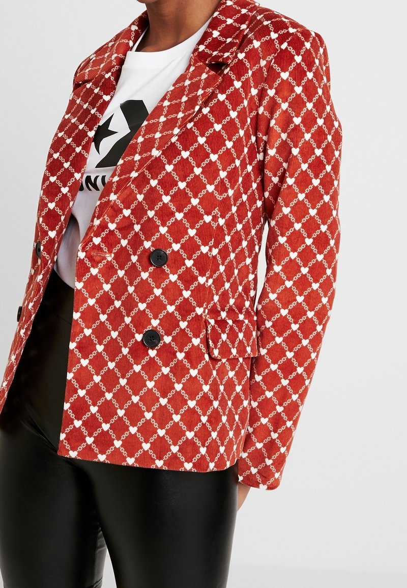 Röd kordfläskjacka med diamantmönster i vitt, dubbelknäppt med svarta knappar och sidofickor, kombinerad med en vit grafisk t-shirt.