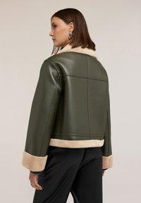 Kobieta zwrócona w lewo, nosząca krótką oliwkowozieloną skórzaną kurtkę z beżowym kołnierzem, mankietami i dolnym brzegiem, w połączeniu z czarnymi spodniami.