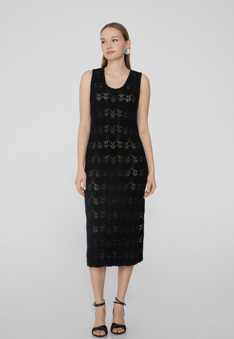Schwarzes ärmelloses Kleid mit einem gestrickten Spitzenmuster, knielang mit einem gewellten Ausschnitt und seitlichen Schlitzern, kombiniert mit schwarzen Sandalen.