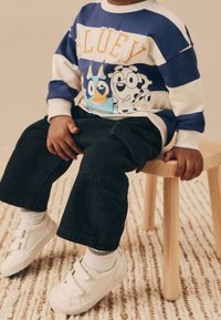Enfant portant un sweat-shirt bleu et blanc à motif de dessin animé, un pantalon noir et des baskets blanches, assis sur un tabouret en bois sur un tapis texturé.