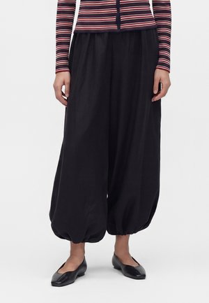 Personne portant un pantalon ballon noir ample avec poignets froncés, des chaussures plates noires, et un haut rayé à manches longues dans des tons rouge, blanc et marine.