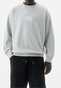 Sweatshirt gris avec un col rond, des poignets et un ourlet côtelés. Présente un imprimé graphique blanc sur la poitrine. Porté avec un pantalon cargo noir.