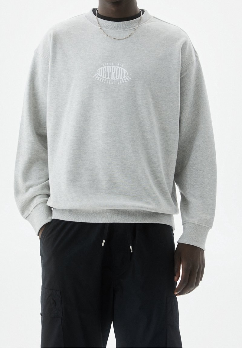 Sweatshirt gris avec un col rond, des poignets et un ourlet côtelés. Présente un imprimé graphique blanc sur la poitrine. Porté avec un pantalon cargo noir.