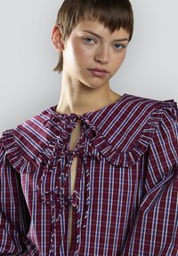 Blusa de cuadros roja y azul con un gran cuello con volantes y lazos en el frente. Presenta mangas largas y un corte holgado con tejido texturizado.