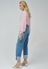 Blusa de color rosa claro con mangas abullonadas, combinada con unos vaqueros de cintura alta en color azul, con los puños doblados. La modelo lleva sandalias de tacón bloque en color beige.
