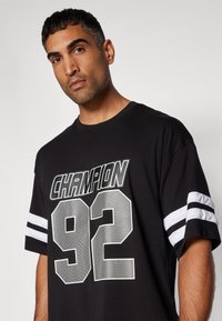 Tricou negru din bumbac cu mâneci cu dungi albe, grafică "Champion" și "92" în model de plasă. Design simplu de tip crew neck.