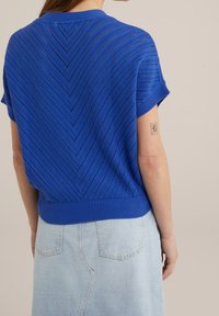 Blå stickad kortärmad topp med ett chevronmönster och ribbad fåll. Bärs tillsammans med en ljusblå denimkjol som syns nedanför.