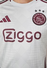 Hvid sportstrøje med maroon accenter, geometrisk mønster, Adidas-logo og Ziggo-tekst. Ajax-emblem med en stiliseret figur og tre stjerner.