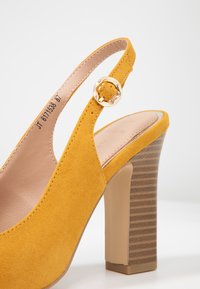 Žluté semišové slingback podpatky s texturovaným dřevěným blokovým podpatkem a zlatou sponou na pásku. Hladká vnitřní podšívka.