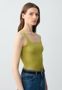 Top verde aderente con scollatura quadrata, abbinato a jeans in denim blu a vita alta e una cintura marrone. Tessuto liscio, design minimalista.