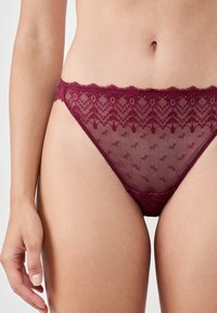 Burgundy lace bikini underwear featuring a scalloped edge, floral pattern, and sheer mesh panels, designed for a fitted silhouette.