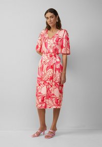 Blumiges Midi-Kleid in sanftem Pink mit roten Mustern, V-Ausschnitt, lockeren Ärmeln und gebundenem Taillengürtel. Kombiniert mit hellrosa Absatzsandalen.