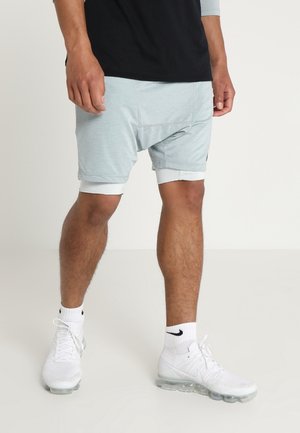 Mann trägt hellgraue, geschichtete Shorts, weiße Nike-Socken und weiße Nike-Sportschuhe und steht auf einem hellen Boden mit einfachem Hintergrund.