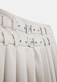 Beige plissee Rock mit silbernen Schnallen und Ösen an doppelten Bändern, glatte Textur und strukturiertes Design, das die Taille betont.