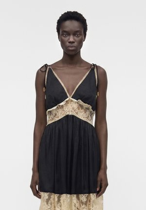 Kvinde med kort krøllet hår iført en ærmeløs sort kjole med beige blonderindsatser og bindebånd, stående foran en ensfarvet baggrund.