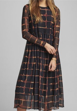 Woman wearing a long-sleeve, black and brown checkered midi dress with sheer sleeves and a gathered waist, standing against a plain background.