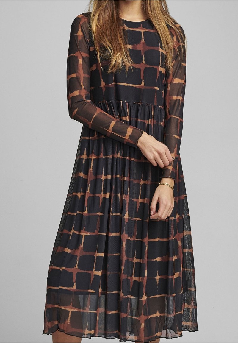 Femme portant une robe midi à manches longues, à carreaux noirs et marron, avec des manches transparentes et une taille froncée, debout devant un fond uni.