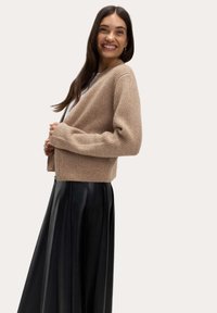 Béžový pletený cardigan s uvolněným střihem, ribovanou texturou a dlouhými rukávy, v kombinaci s černou skládací sukní. Hladká tkanina a zkrácená délka.