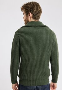 Pull à côtes vert foncé avec un large col, des manches longues et un tricot texturé. Présente une taille cintrée et est porté avec un jean sombre.