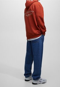 Sudadera de mezcla de algodón en naranja quemado con letras blancas, combinada con jeans azules holgados y zapatillas grises. Ajuste casual y relajado.