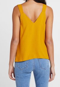 Donna che indossa un top senza maniche di colore giallo senape con schiena a V e jeans in denim blu chiaro, in piedi davanti a uno sfondo chiaro e uniforme.