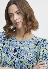 Blumenbluse mit einem mehrfarbigen Muster aus blauen und grünen Blumen auf einem cremefarbenen Hintergrund. Strukturierter Stoff mit rundem Ausschnitt.