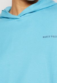 Hoodie bleu clair en tissu doux. Doté d'une capuche avec cordon de serrage et d'un logo imprimé en bleu foncé sur la poitrine. Design simple et décontracté.